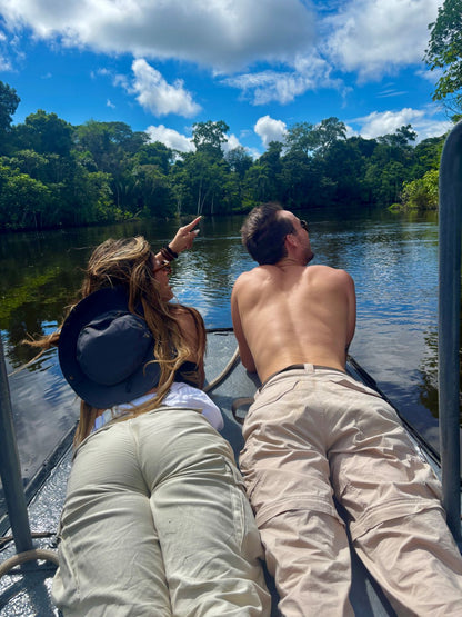 Two people in a boat on the amazon  river surrounded by lush greenery under a blue sky with clouds.