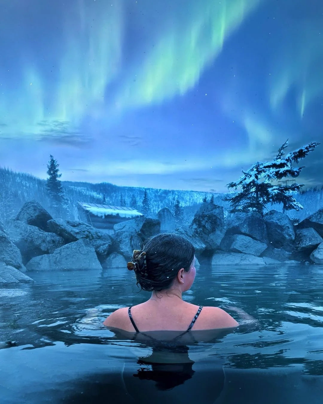 Person in a hot tub under the Northern Lights with snowy landscape